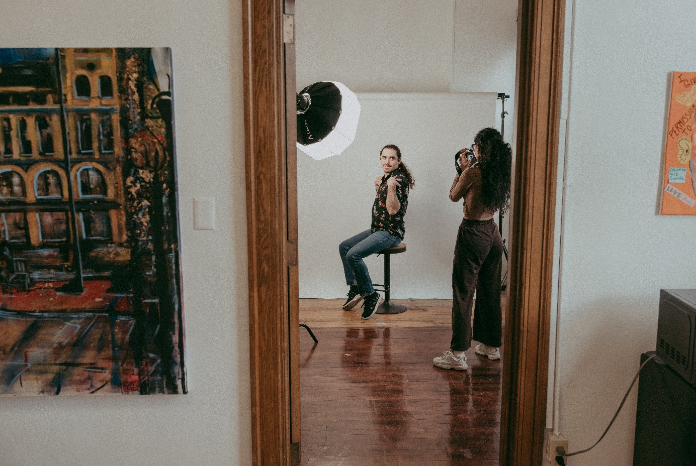 A woman stands inside of a doorway taking a photo of a person sitting on a stool.