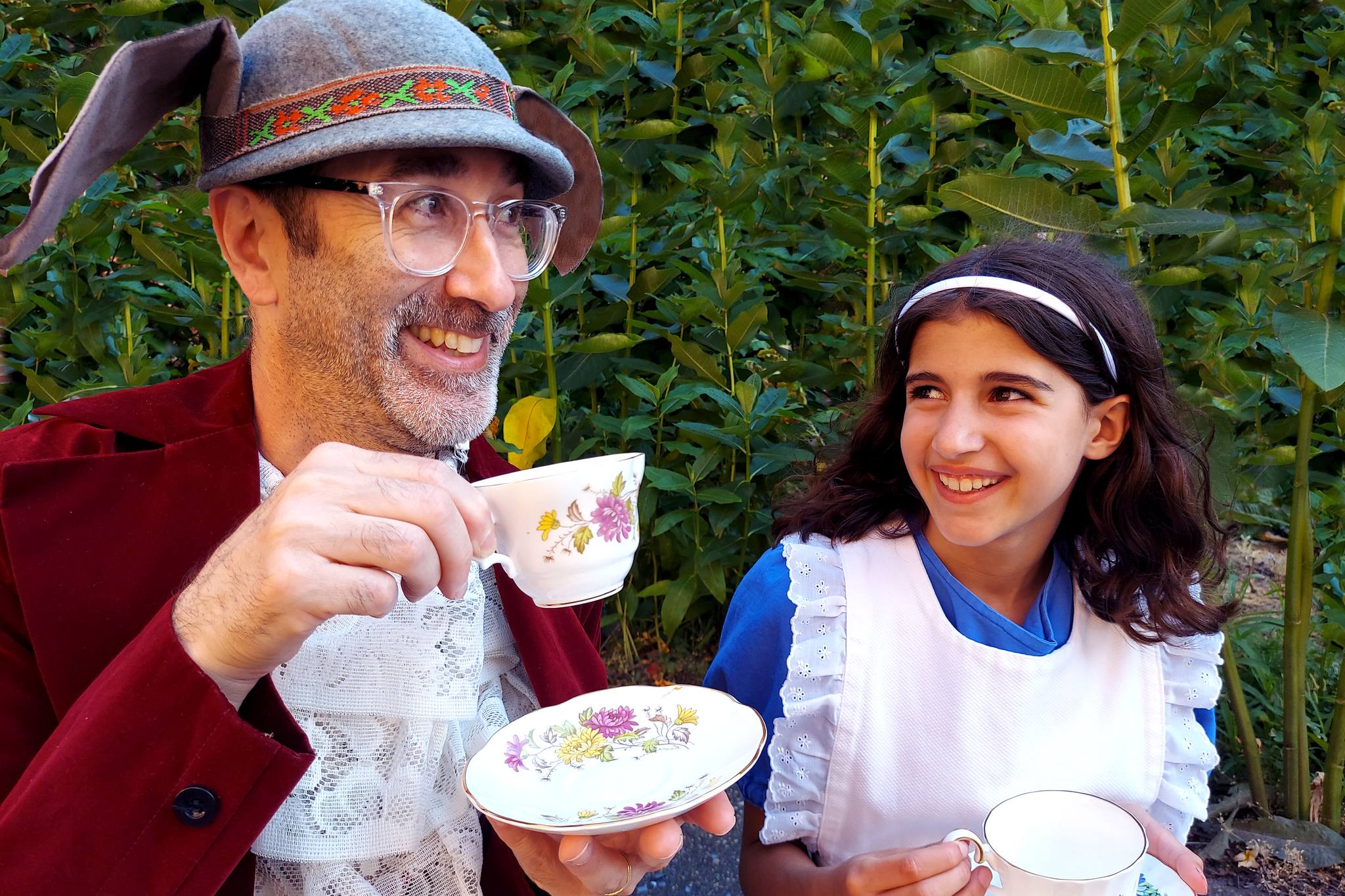 A man wearing a rabbit hat drinks tea with a girl dressed as Alice in Wonderland